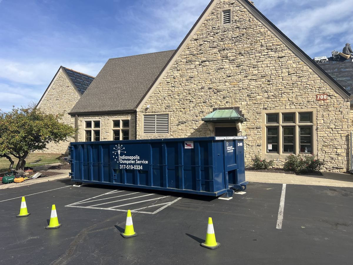 A navy Indianapolis Dumpster Service roll off container. In a parking lot beside a stone building.