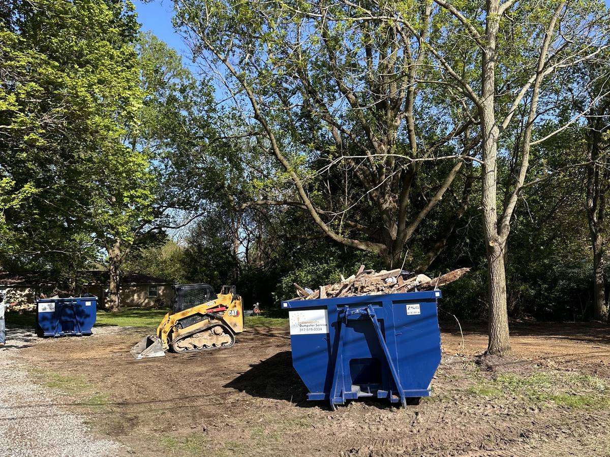 A navy Indianapolis Dumpster Service roll off container. On a cleared lot, with a skid steer at work.