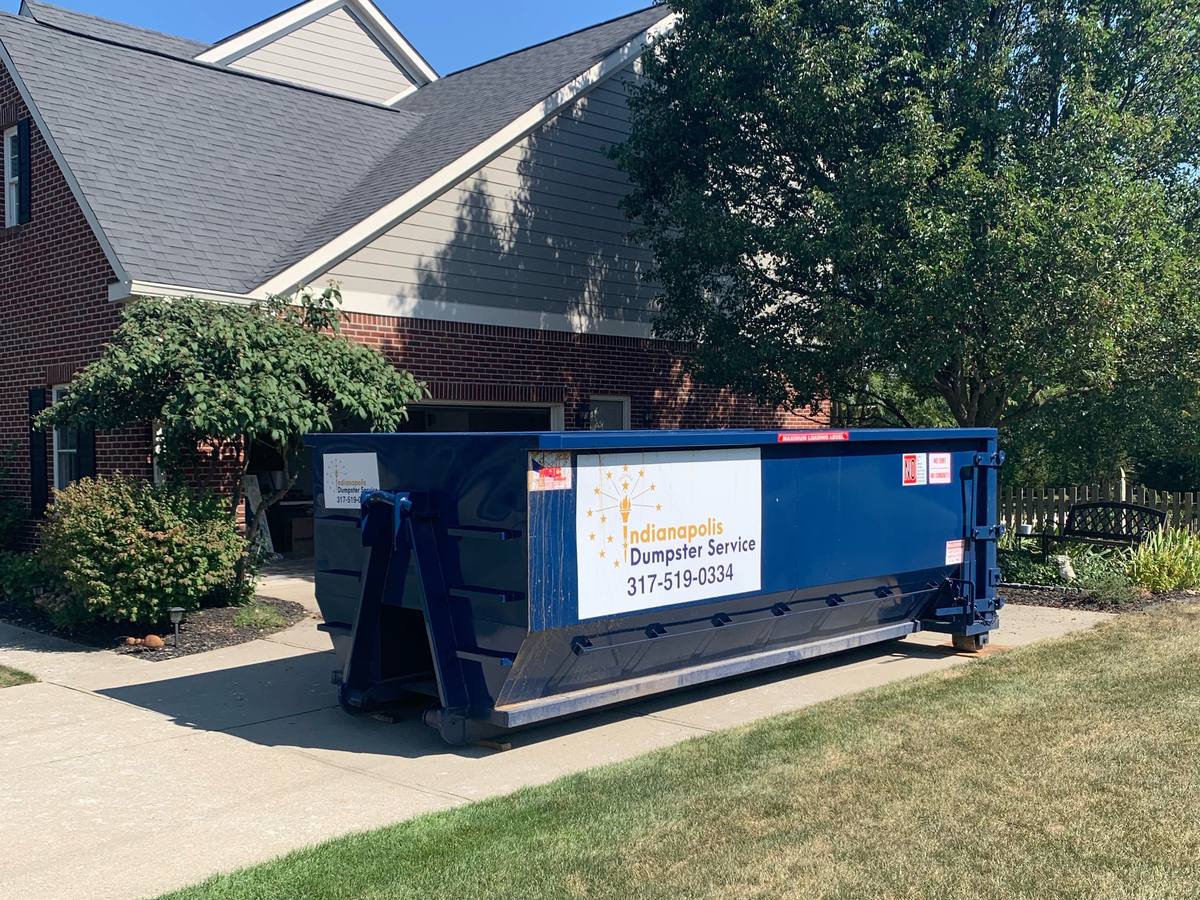 A navy Indianapolis Dumpster Service roll off container. On the driveway of a brick home.