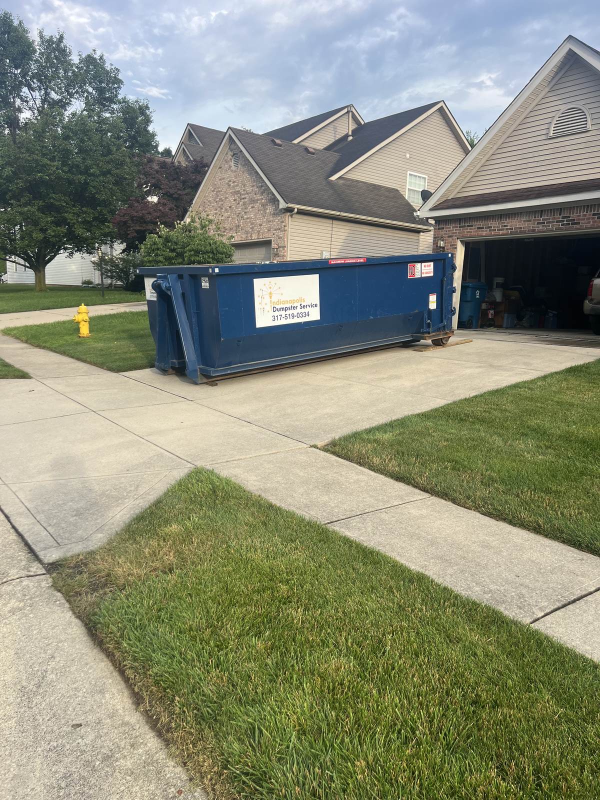 A navy Indianapolis Dumpster Service roll off container in Carmel. In front of the garage on a brick-front home.