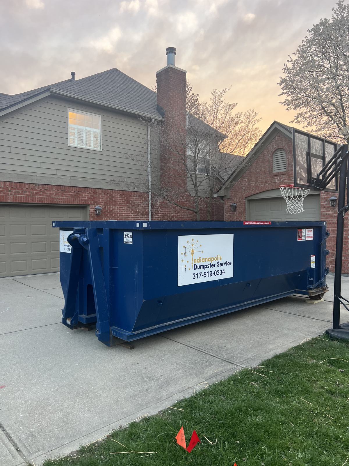 A navy Indianapolis Dumpster Service roll off container in Carmel. Beside a brick home in the evening.