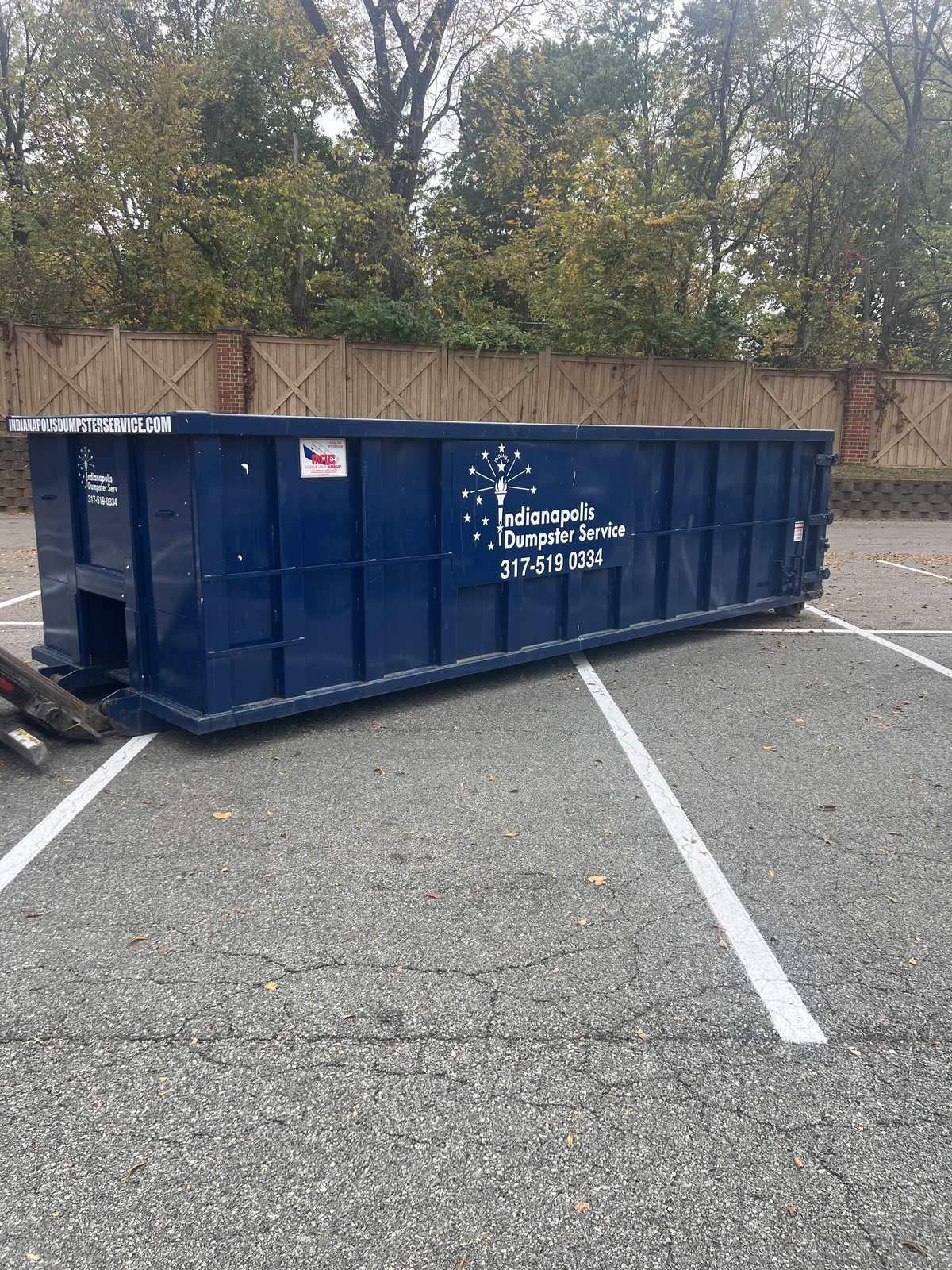 A navy Indianapolis Dumpster Service roll off container in Carmel. Parked across the spaces of a fenced lot.