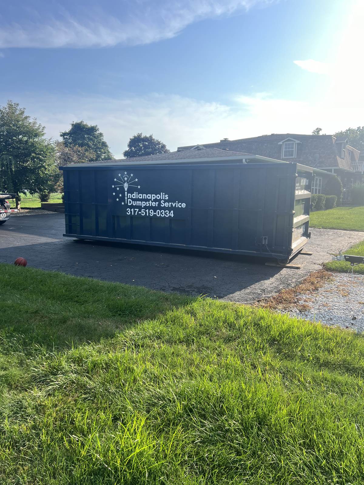 A navy Indianapolis Dumpster Service roll off container in Cicero. On a blacktop drive at the edge of the lawn.