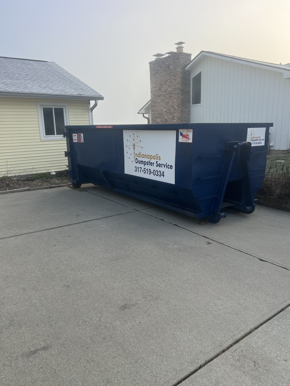 A navy Indianapolis Dumpster Service roll off container in Cicero. On a concrete driveway beside a two-story home.