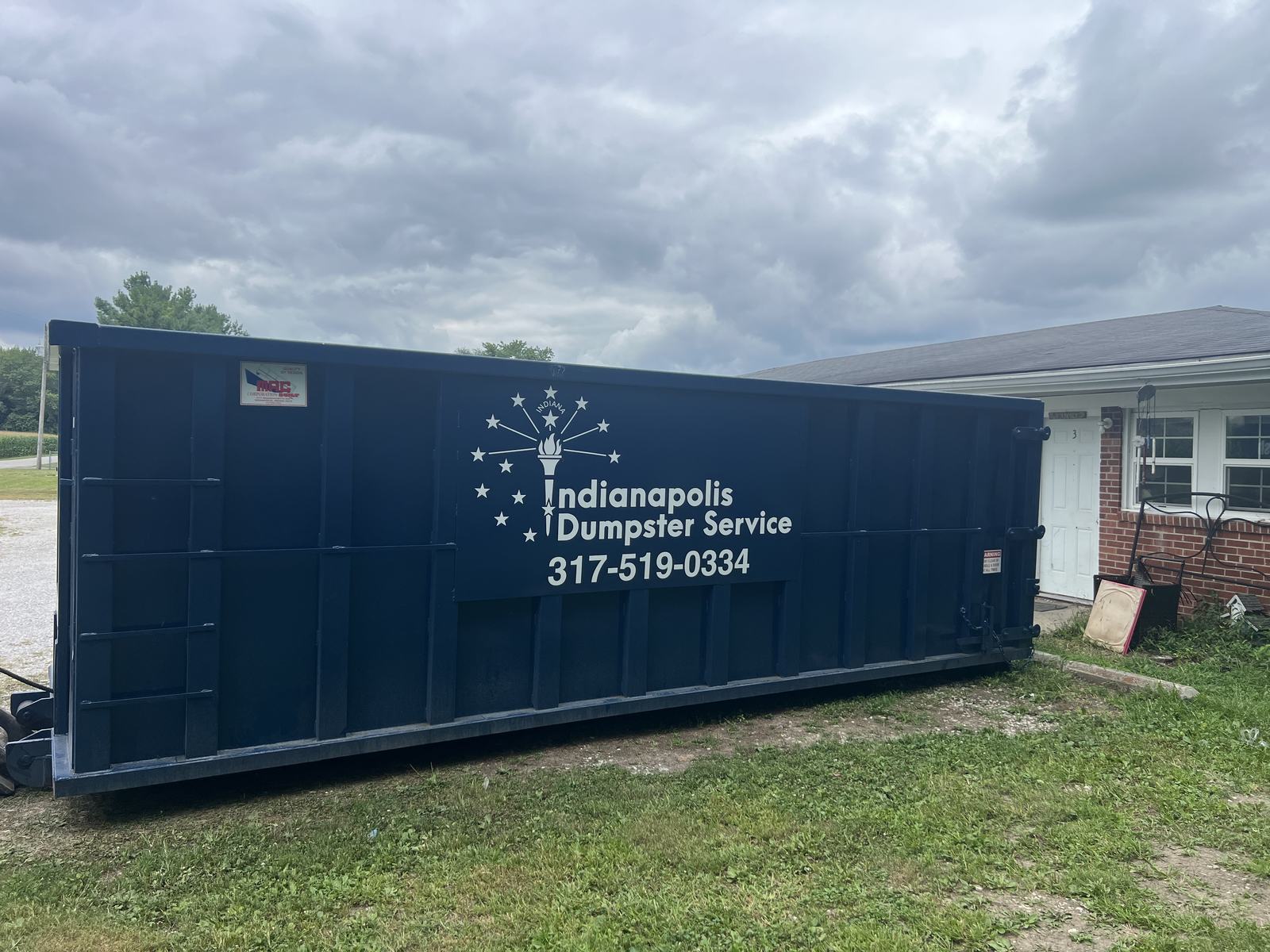 A navy Indianapolis Dumpster Service roll off container in Elwood. In the side yard of a brick ranch home.