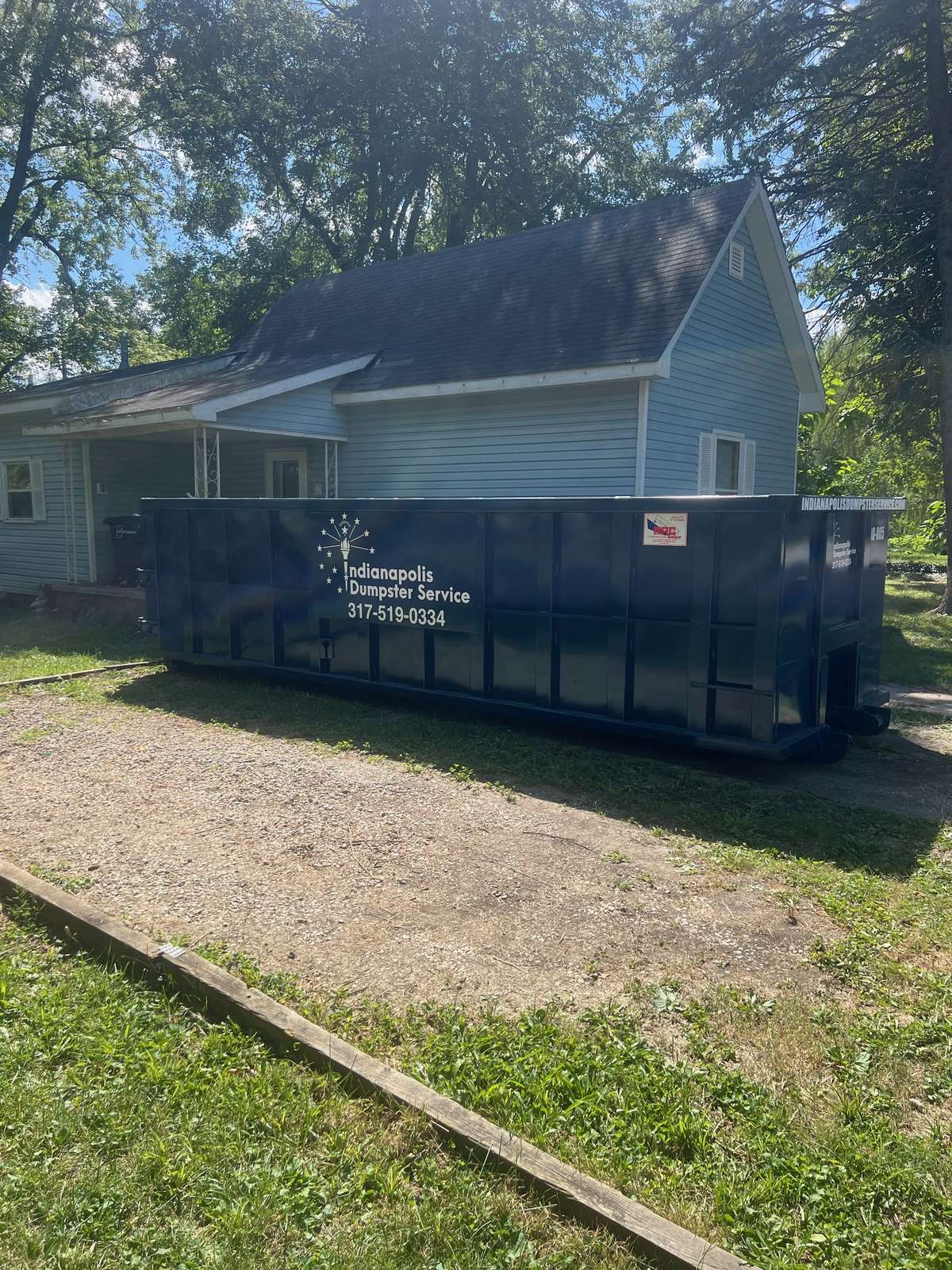 A navy Indianapolis Dumpster Service roll off container in Elwood. On a gravel pad beside a blue house.
