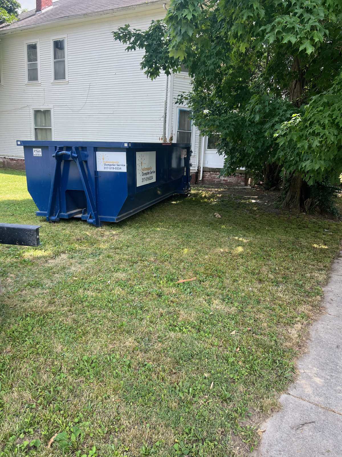 A navy Indianapolis Dumpster Service roll off container in Indianapolis. Against the garage apron of a white-sided home.