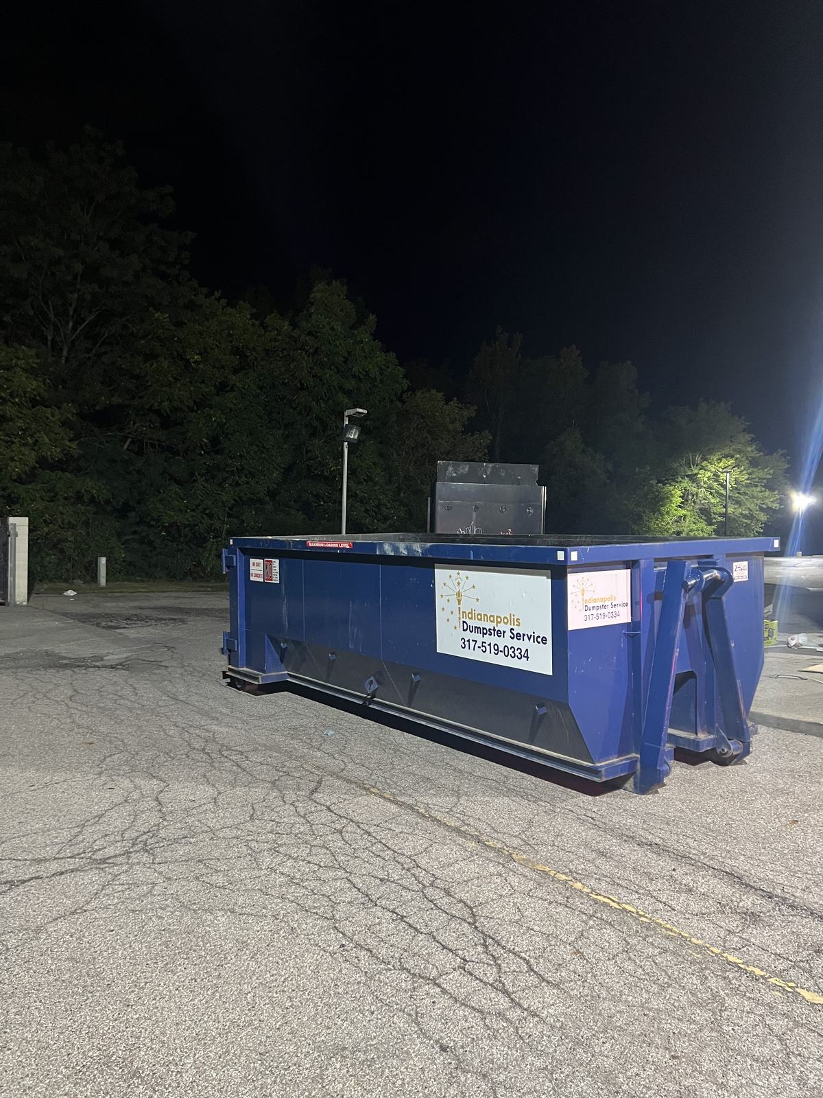 A navy Indianapolis Dumpster Service roll off container. In an empty parking lot after dark.