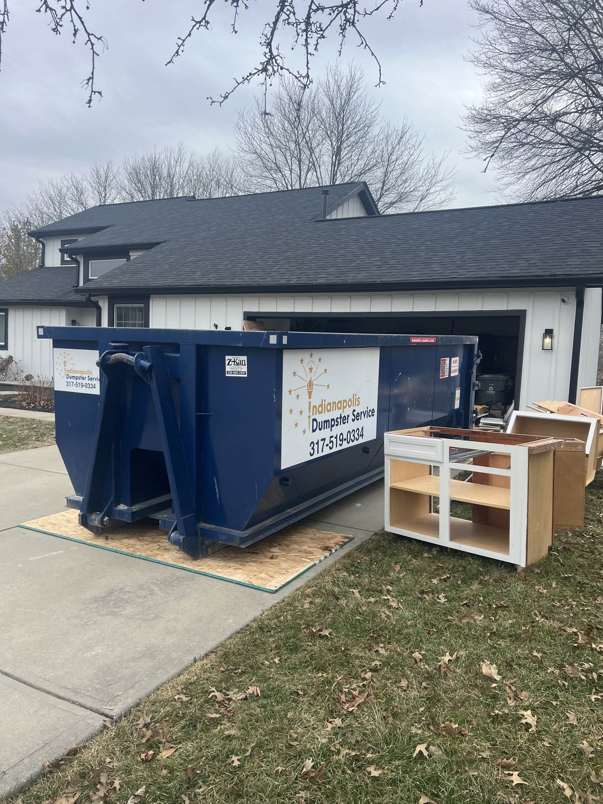 A navy Indianapolis Dumpster Service roll off container in Noblesville. Old kitchen cabinets stacked beside the dumpster.