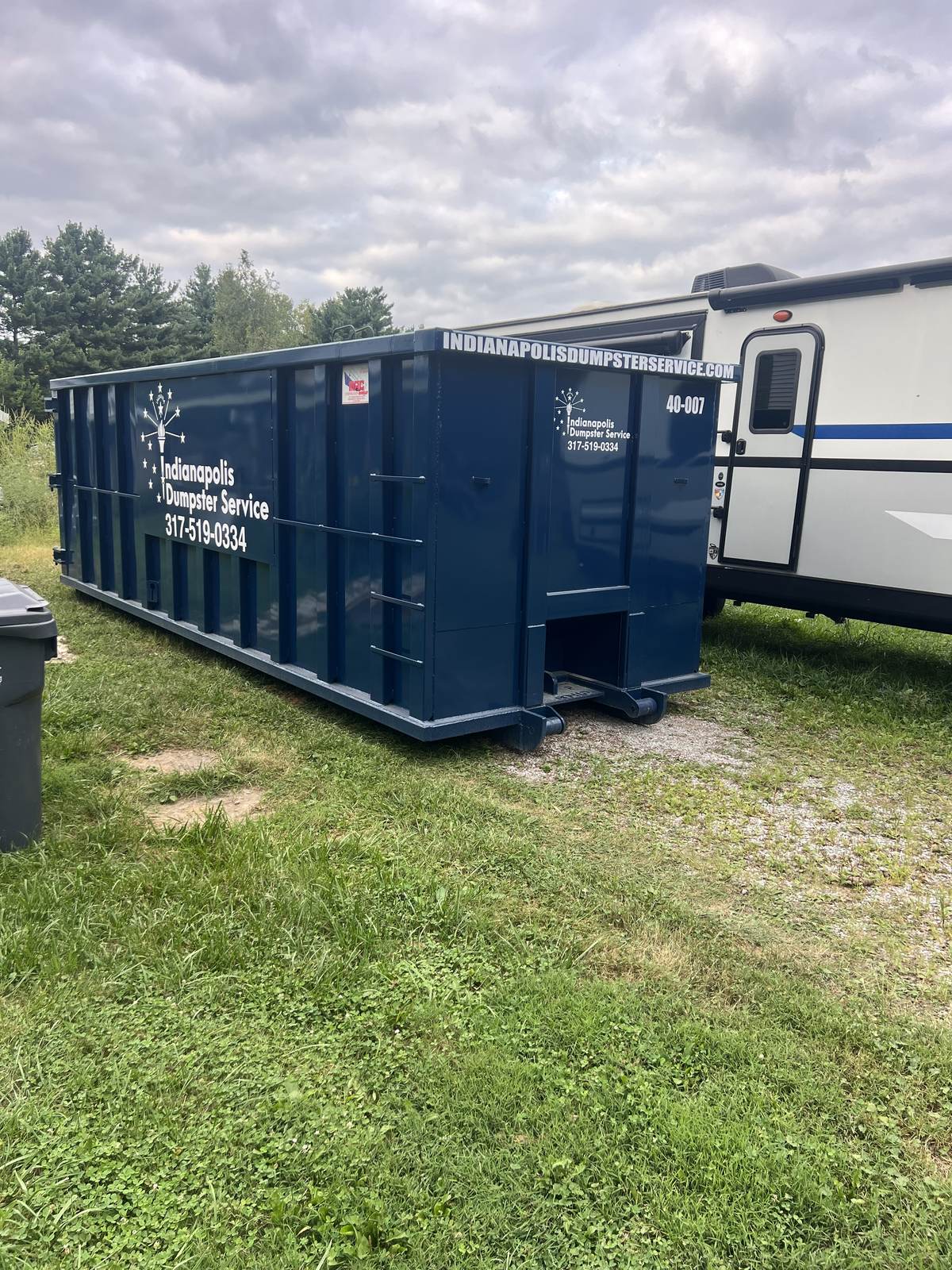 A navy Indianapolis Dumpster Service roll off container in Pendleton. On the grass next to a parked camper.