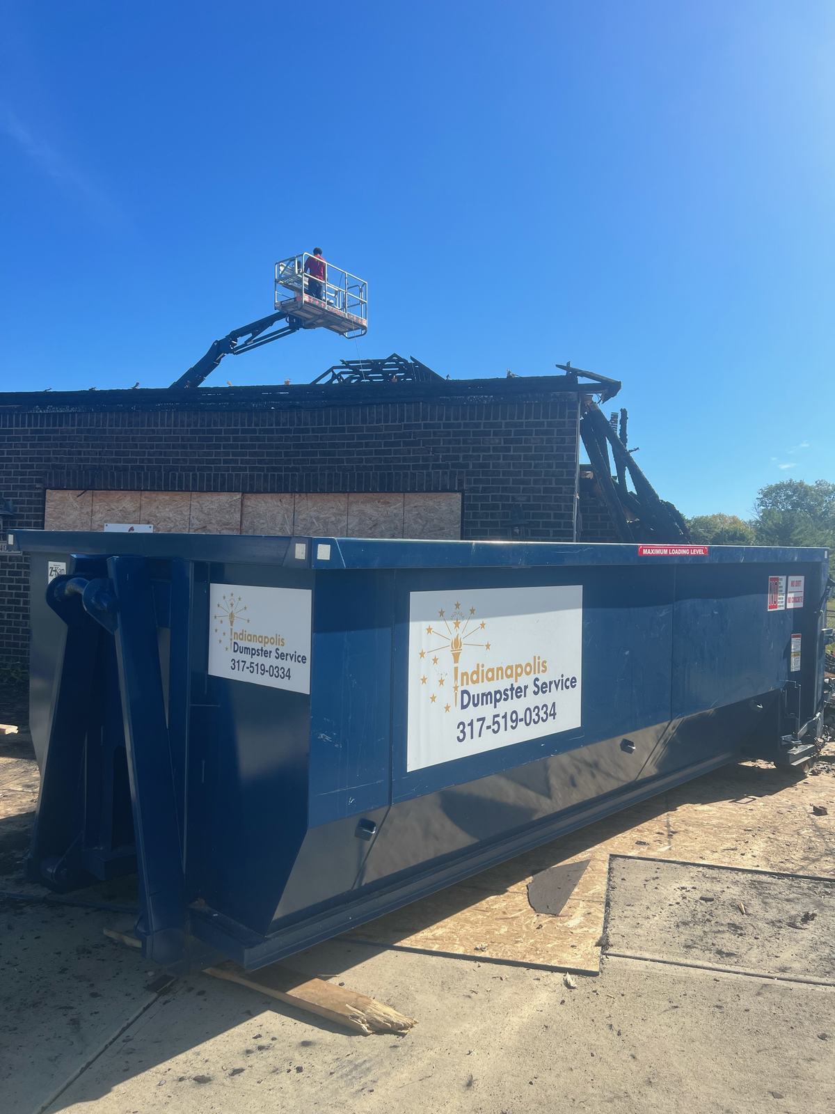 A navy Indianapolis Dumpster Service roll off container. A lift at the roofline with the dumpster below.
