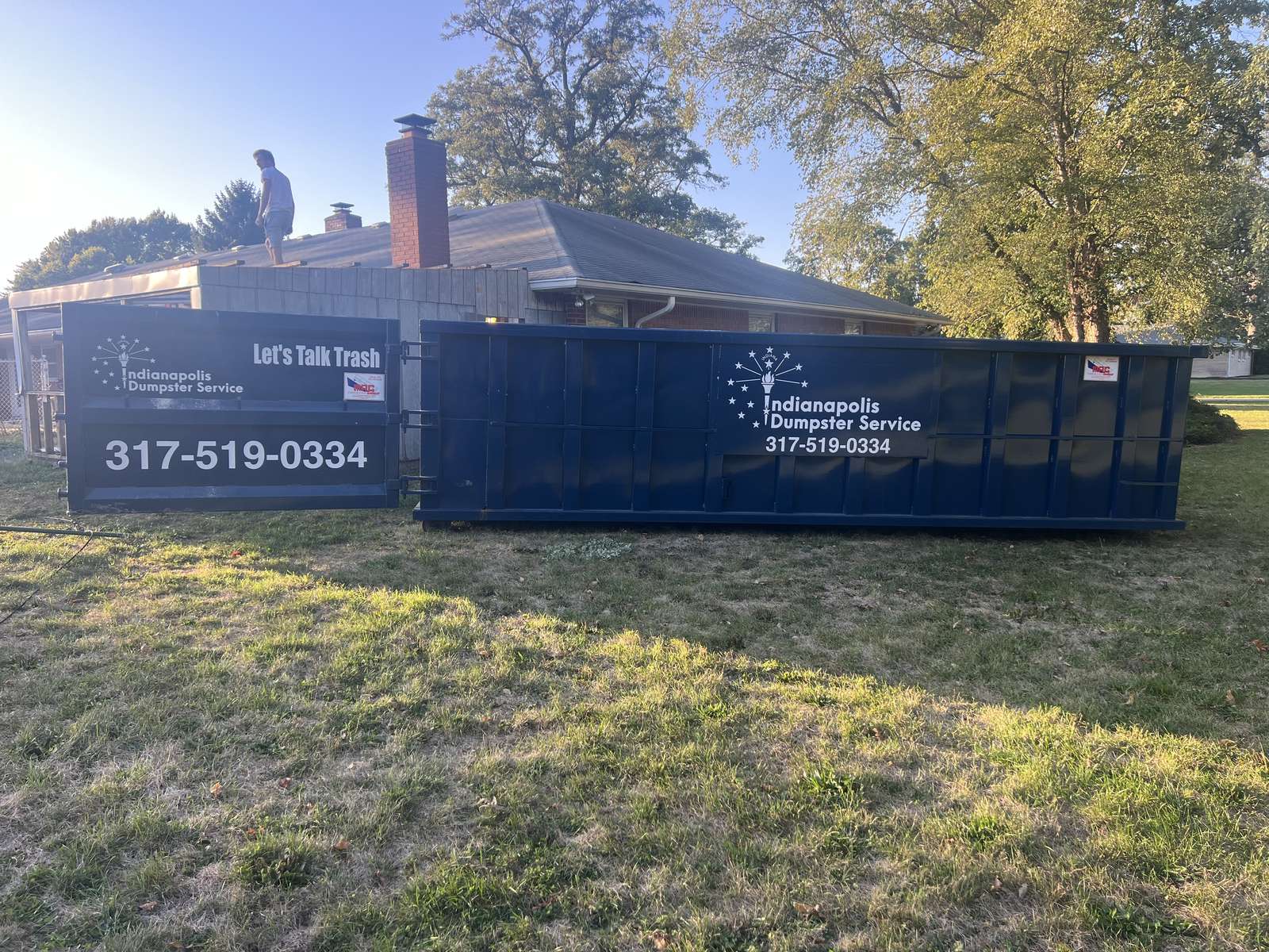 A navy Indianapolis Dumpster Service roll off container in Sheridan. On the lawn while a roofer works on the house.